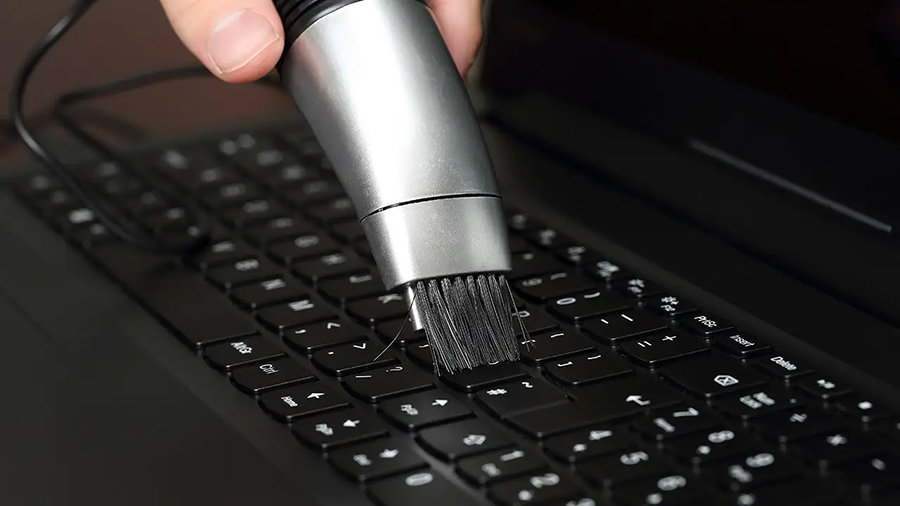 A laptop's keyboard is being cleaned by someone using a brush attachment on a vacuum cleaner.